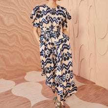 Load image into Gallery viewer, The Marion Dress boasts a striking ikat motif with electric blue graphics against a peach background. This short sleeve dress has flattering ties at the waist and a ruffled peplum before cascading to an elegant midi hemline. It has a button keyhole closure at the back.   100% Viscose
